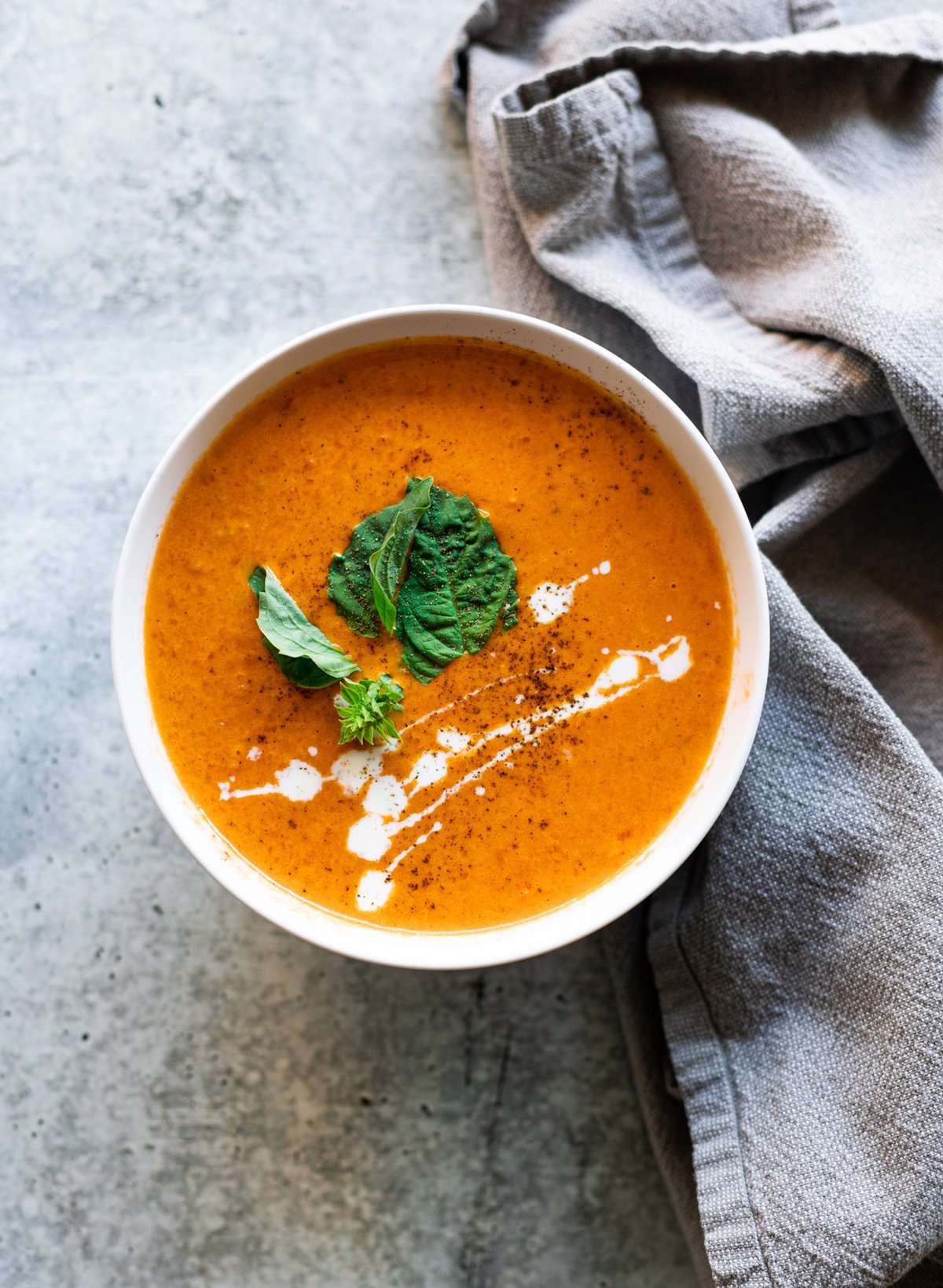 A bowl of roasted red pepper soup garnished with fresh basil, a drizzle of heavy whipping cream, and freshly cracked black pepper. 