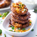Stack of corn fritters on a platter topped with shredded cheese and bacon.